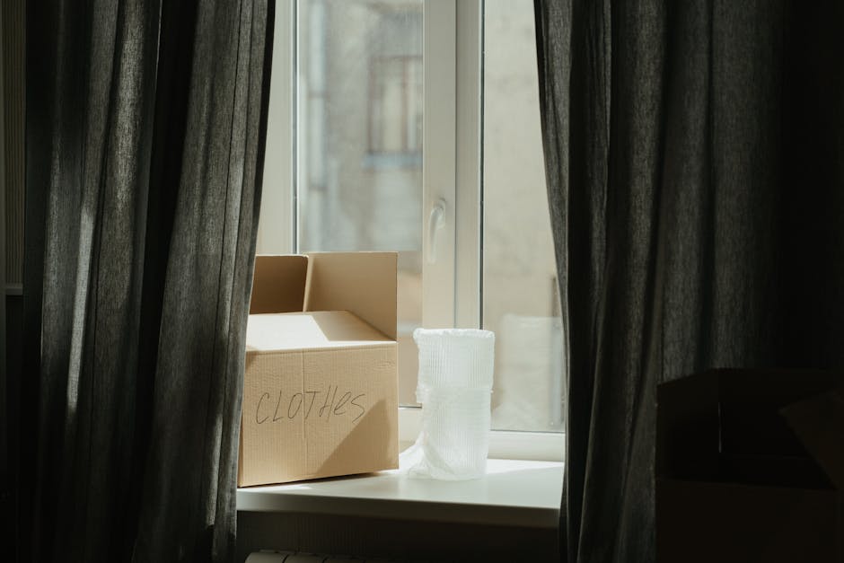 A cardboard box labeled 'Clothes' is placed on a white windowsill inside a home during packing for a house move, with rolled-up white fabric or clothing next to it. The window is partially covered by 