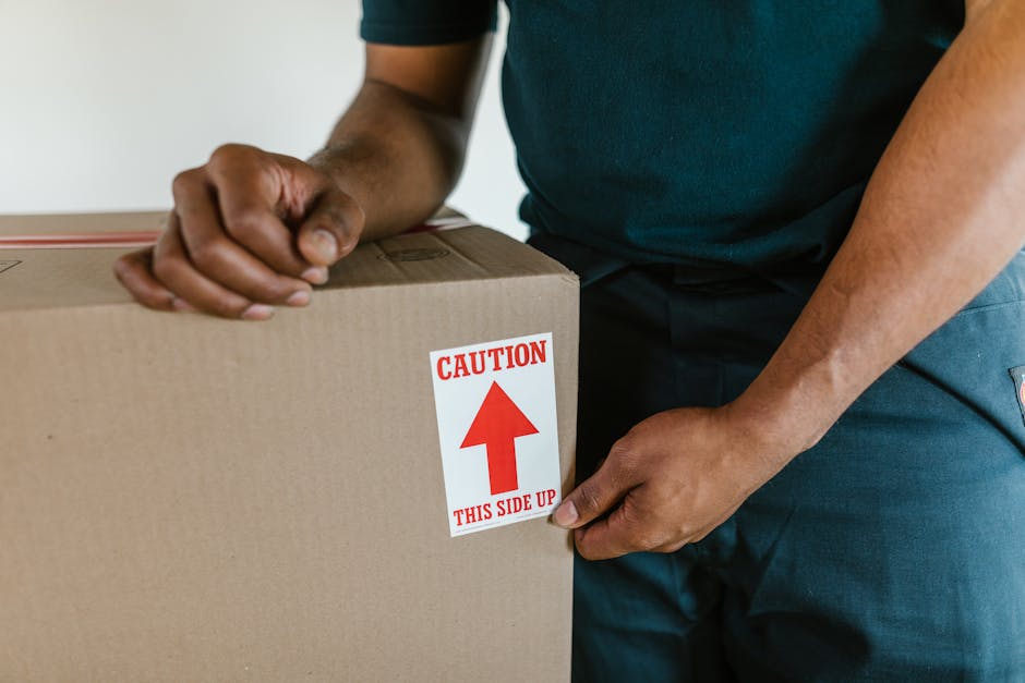 A cardboard box labeled 'Clothes' is placed on a white windowsill inside a home during packing for a house move, with rolled-up white fabric or clothing next to it. The window is partially covered by 