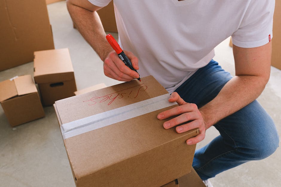 A person wearing a white t-shirt and blue jeans is crouched down, using a red marker to write the word 'Specs' on a medium-sized cardboard box secured with white packing tape, inside a room surrounded