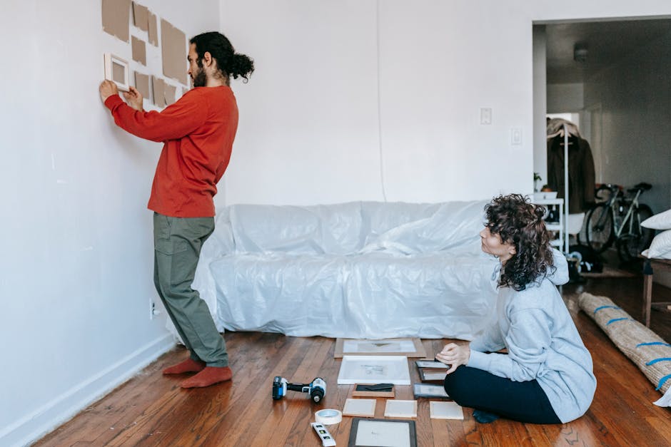A person with a red sweater and grey trousers is kneeling on a wooden floor, holding a small wooden picture frame as part of a home relocation process. A woman with curly dark hair, wearing a grey hoo